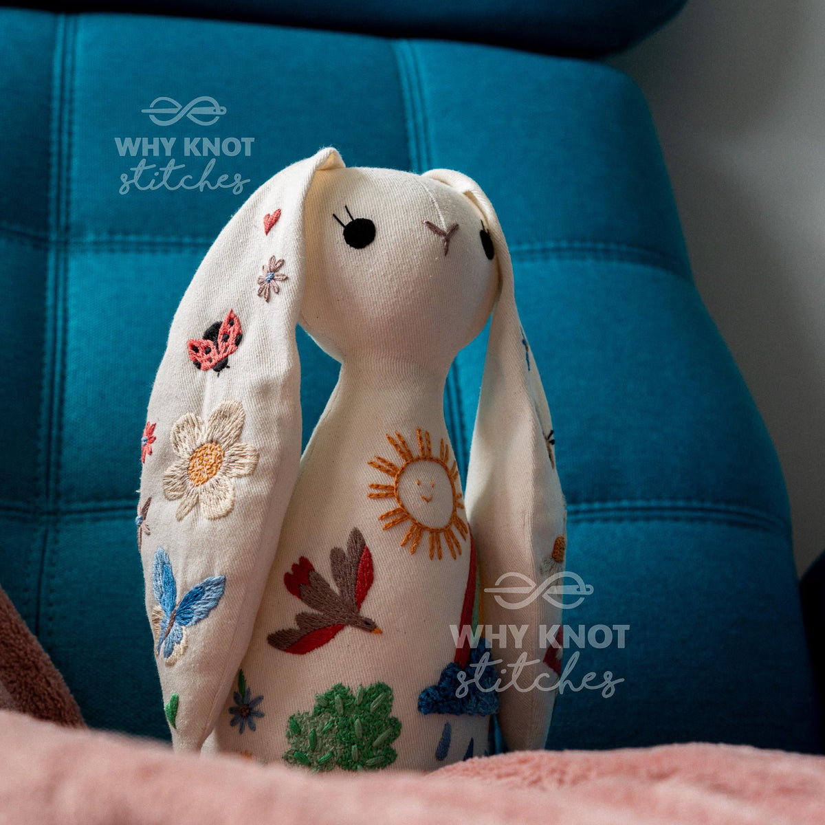 May include: An off-white fabric rabbit plush toy featuring embroidered designs including a sun, a bird, a butterfly, a ladybug, a flower, a cloud, and a rainbow. The rabbit has long ears, black circular eyes, and simple stitched facial features.