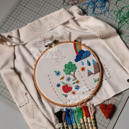 May include: A round wooden embroidery hoop containing off-white fabric with colorful stitched designs of a tree, apple, flowers, mountains, and a rainbow cloud. The fabric features printed outlines and is accompanied by several embroidery floss skei