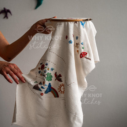 May include: A round wooden embroidery hoop holding a piece of off-white fabric with colorful embroidered designs including a red mushroom, a rainbow, a sun, flowers, and a tree. The text WHY KNOT stitches is printed on the fabric in white.
