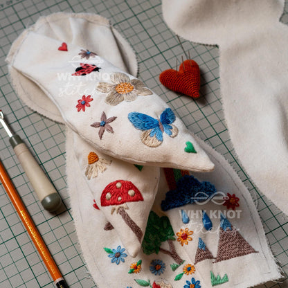 May include: Off-white fabric pieces featuring colorful embroidery of a red mushroom, blue butterfly, ladybug, flowers, mountains, and a rain cloud. A red heart-shaped pincushion, a seam ripper, and an orange pencil are placed nearby on a green grid 