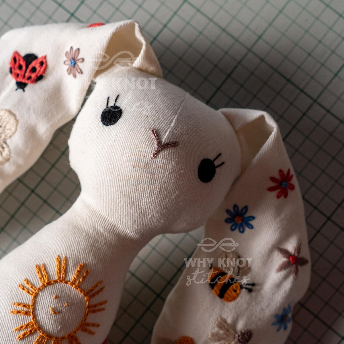 May include: A cream-colored fabric rabbit plush toy featuring embroidered details including a yellow sun, a red ladybug, a bee, and various colorful flowers. The rabbit has black stitched eyes and a brown stitched nose, with the text Why Knot Stitch