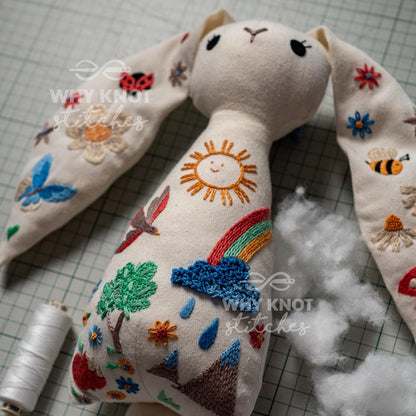 May include: An off-white fabric rabbit doll featuring colorful hand-embroidered designs including a sun, rainbow, rain cloud, mountains, trees, flowers, a butterfly, a ladybug, and a bee. The text "WHY KNOT STITCHES" is printed over the doll.
