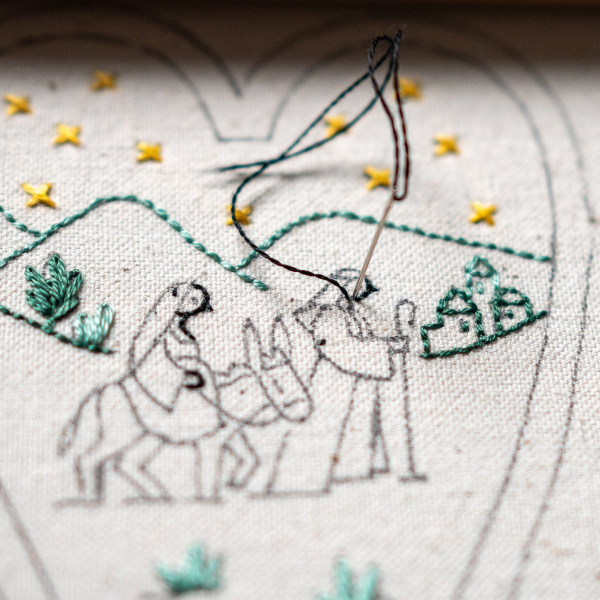 Hand embroidery in progress showing the outlined figures of Mary, Joseph, and the donkey beneath embroidered golden stars and green hills.