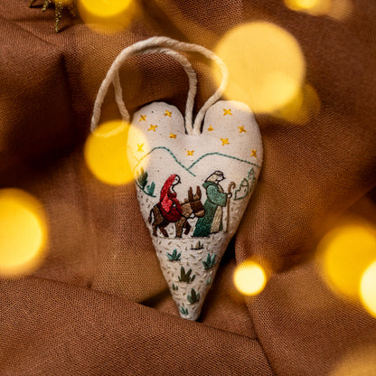 Handmade nativity heart ornament hanging by its cotton cord against warm brown fabric, surrounded by soft golden Christmas lights.