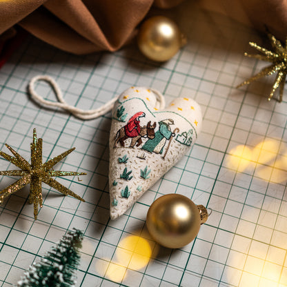 Finished Road to Bethlehem embroidered heart ornament with a cotton hanging loop, styled with golden Christmas decorations and festive lights.