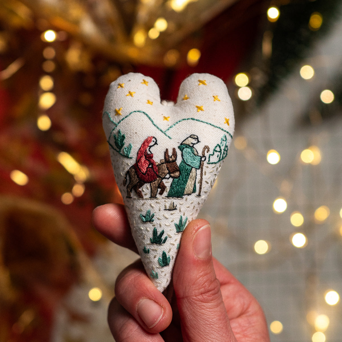 Hand-embroidered Christmas heart ornament featuring Mary riding a donkey alongside Joseph on the road to Bethlehem, with golden stars and a softly lit festive background.
