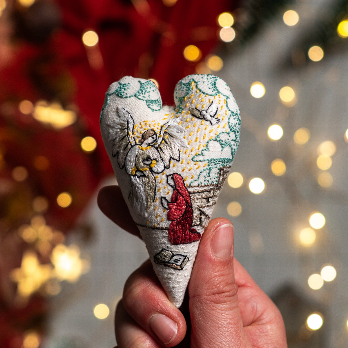 The Annunciation handmade embroidered heart ornament featuring Angel Gabriel and Mary, held against warm Christmas lights.